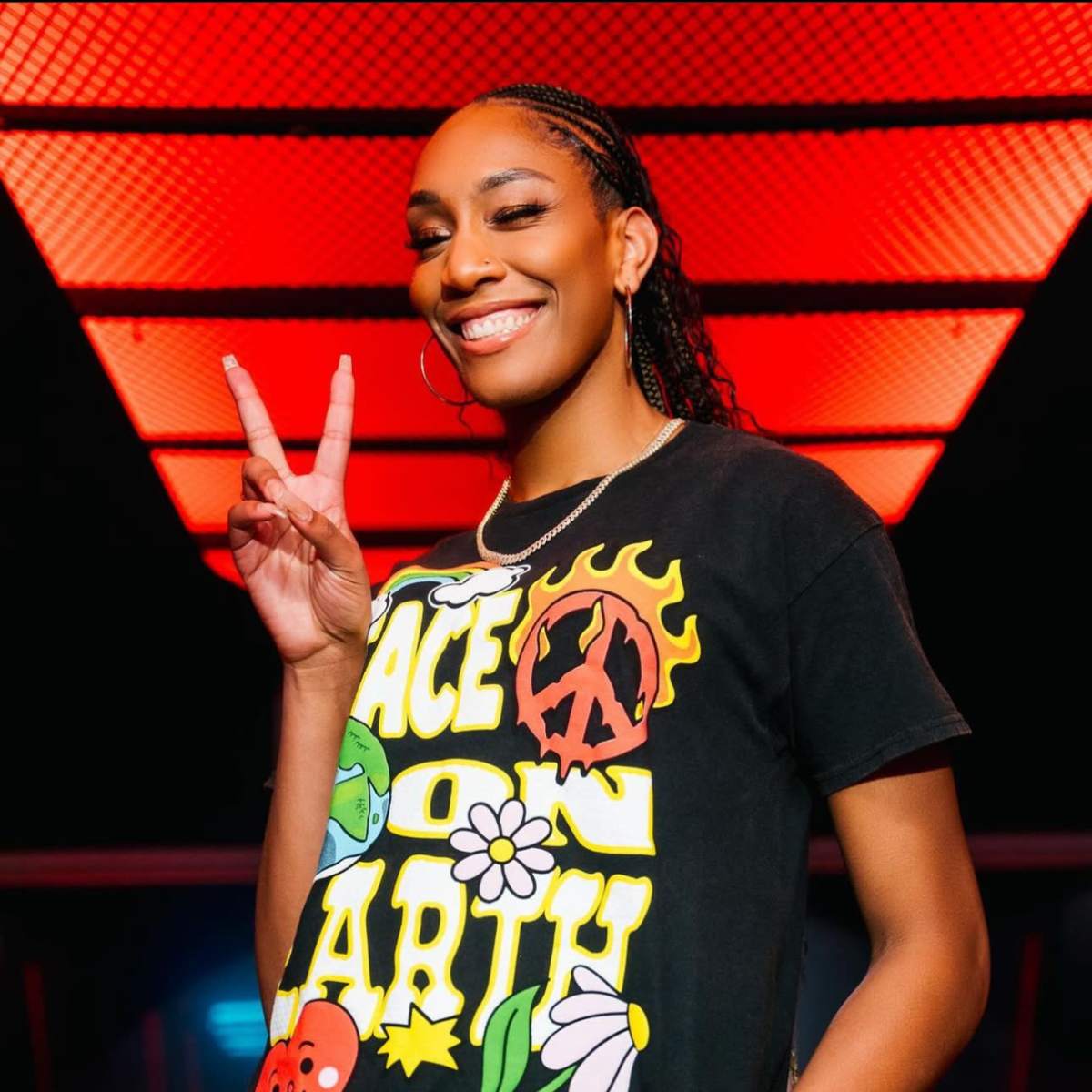 A person wearing a colorful graphic t-shirt poses with a peace sign against a backdrop of red lighting.