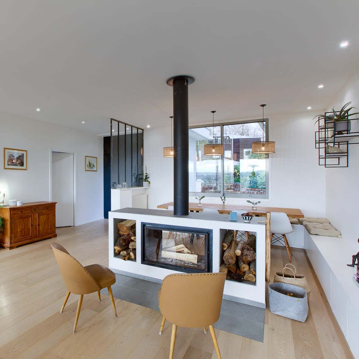 A modern open-concept living space that features a central fireplace, a dining area with a large window, and a staircase in the background.