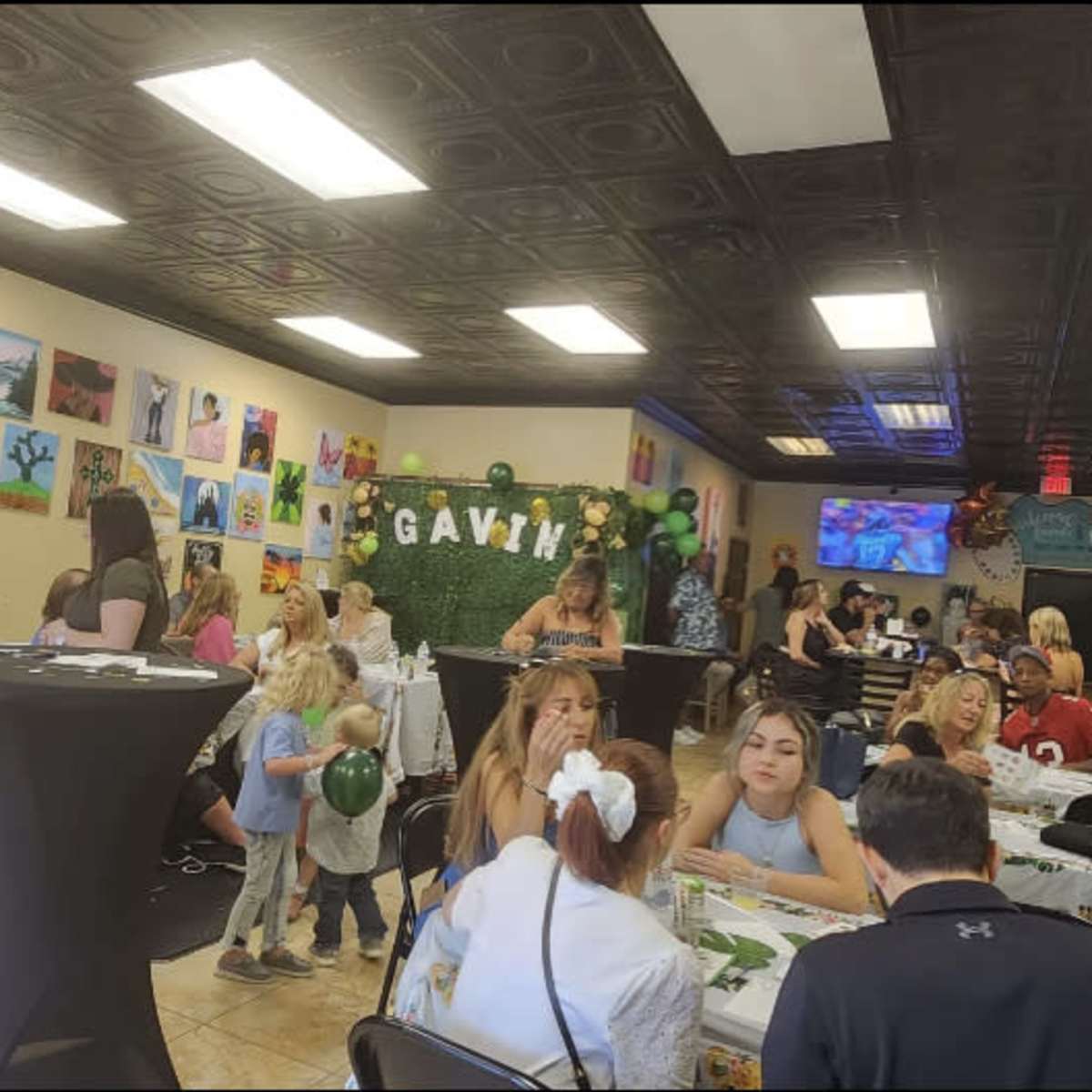 A lively indoor event with guests seated at tables decorated with festive tablecloths, surrounded by artwork on the walls and balloons.