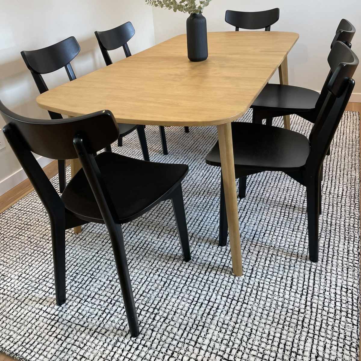 A wooden dining table surrounded by six black chairs, with a simple vase holding greenery in the center, placed on a textured rug.