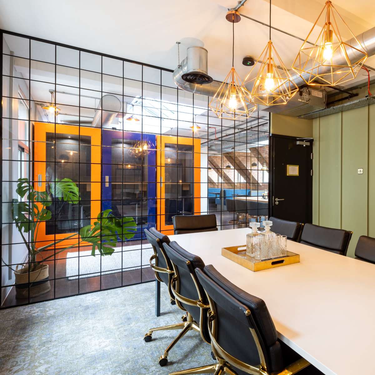 A modern conference room features a large white table surrounded by black chairs, with geometric pendant lights and a glass partition leading to a space with orange doors.