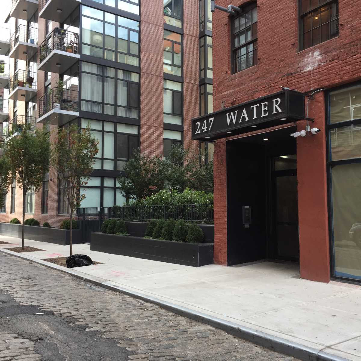 A street scene featuring a red brick building with a sign that says "247 WATER" and modern apartment buildings in the background.
