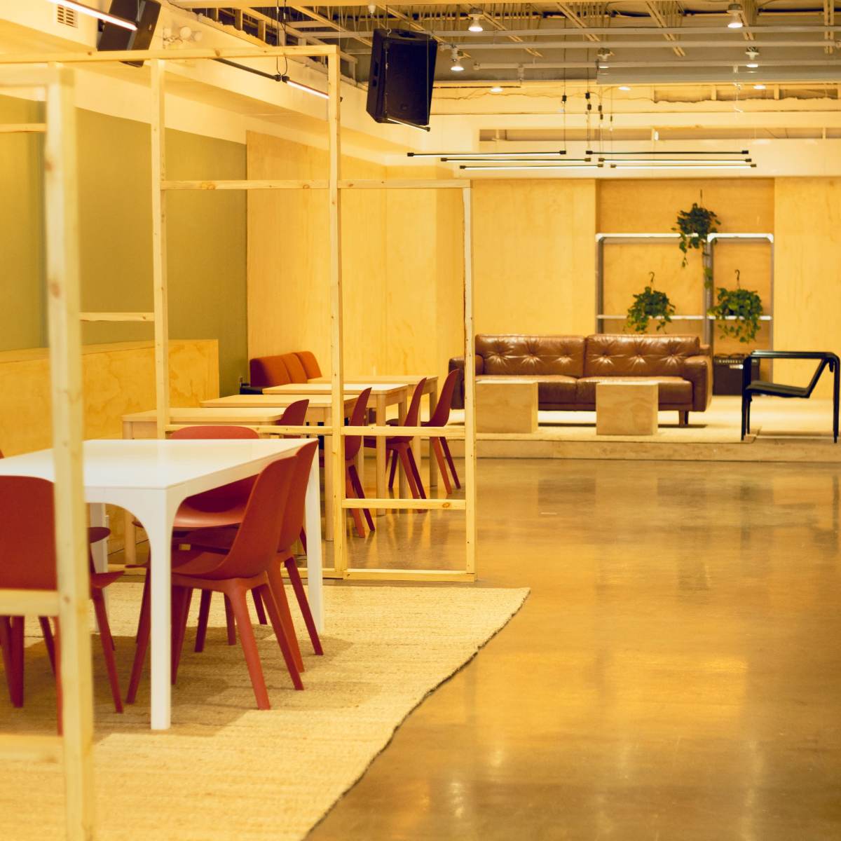 A modern, open workspace featuring wooden structural frames, red chairs around white tables, and a seating area with brown couches and plants.