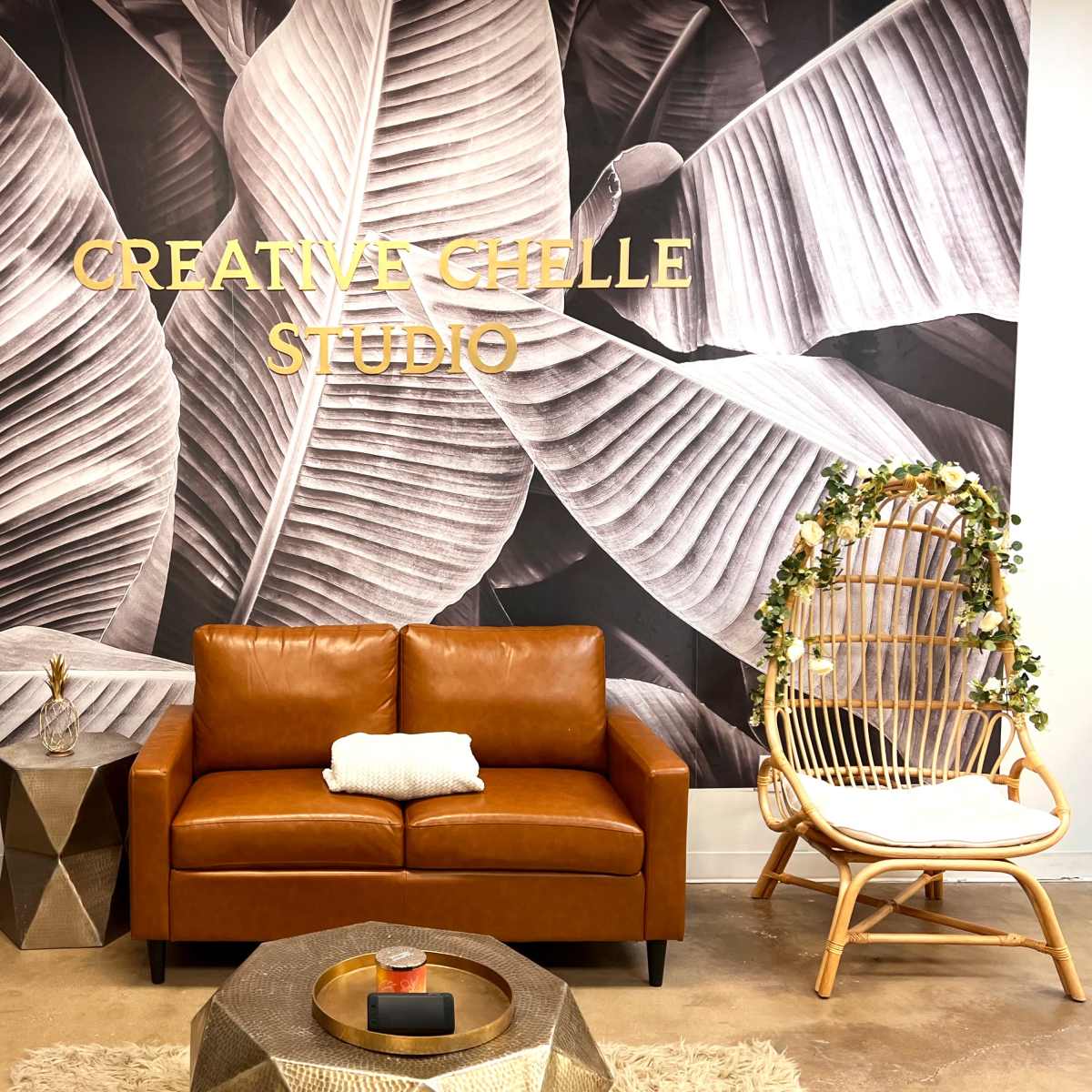 A cozy studio space with a brown leather sofa, a decorative gold side table, and a rattan chair surrounded by a wall featuring large leaf patterns and the words "Creative Chelle Studio."