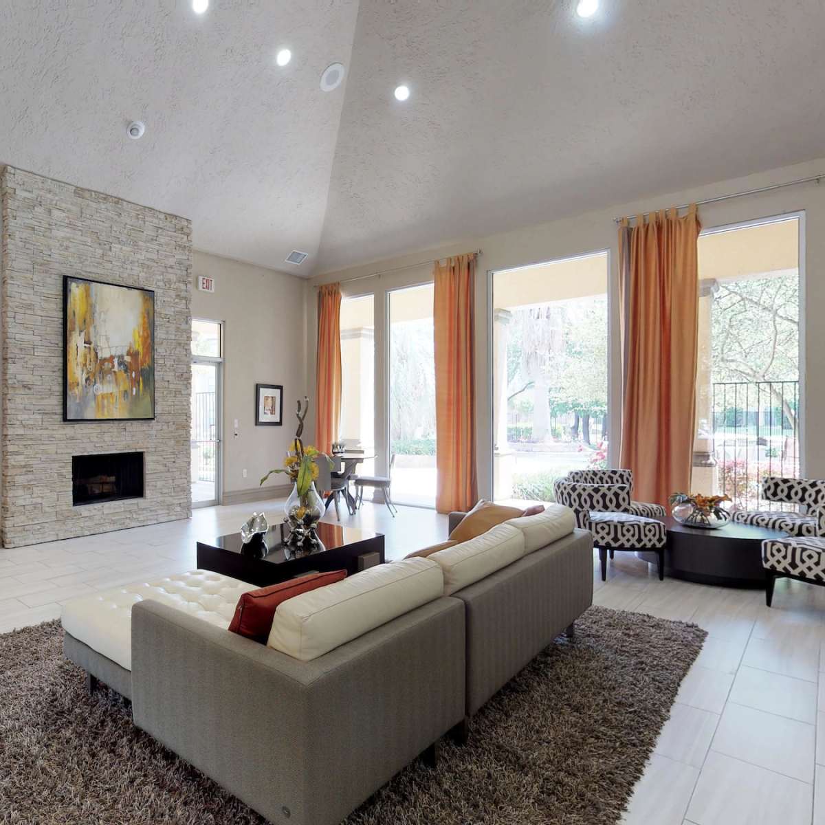 A modern living room with a stone fireplace, a beige sofa, a coffee table, and large windows draped with orange curtains.
