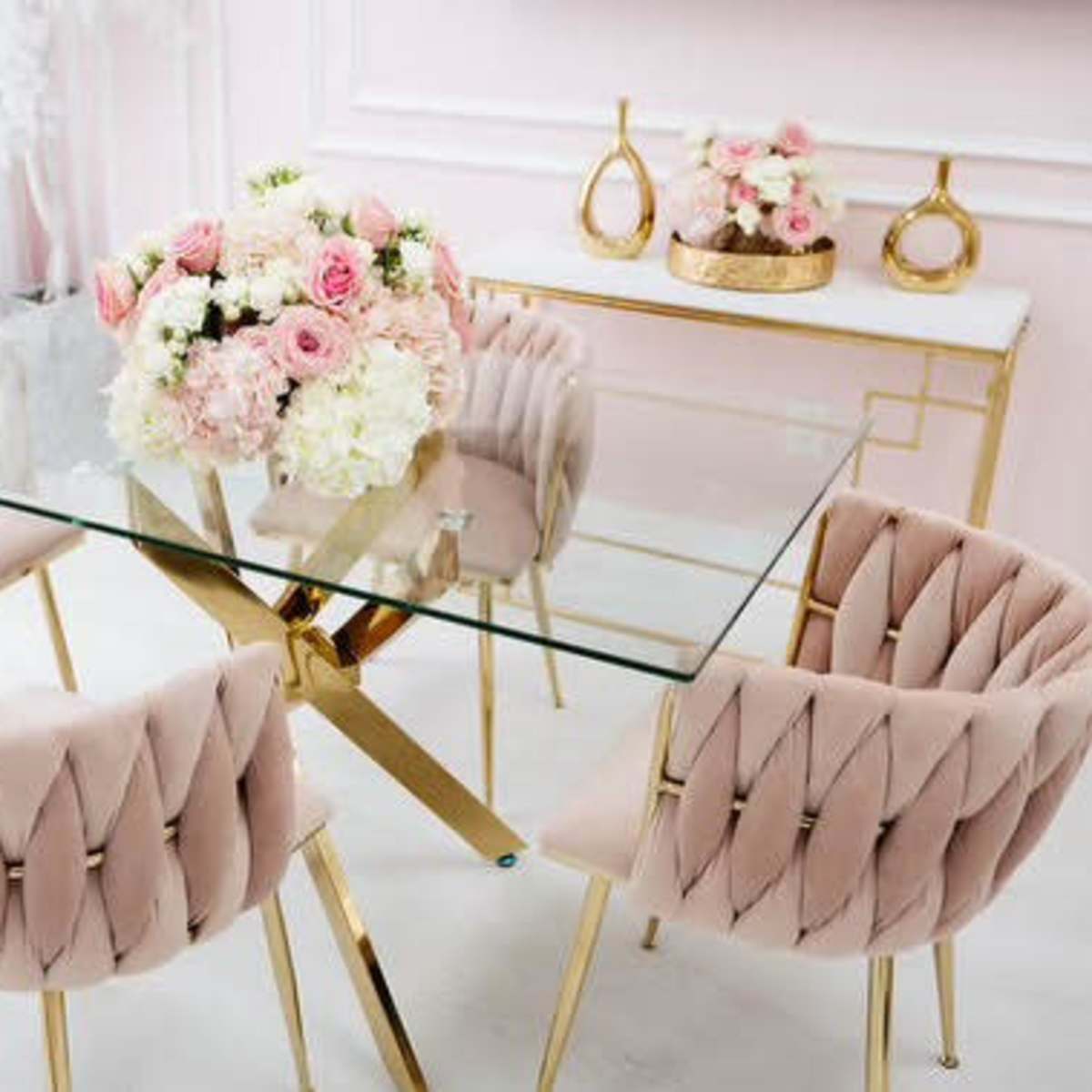 A glass dining table surrounded by four pink upholstered chairs is set in a room with light pink walls and decorative floral arrangements.