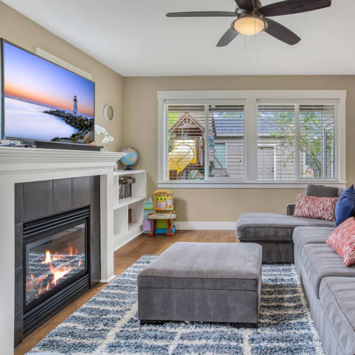 The living room features a gray sectional sofa, a television mounted above a fireplace, and a dining area with modern chairs and a visible play structure outside.