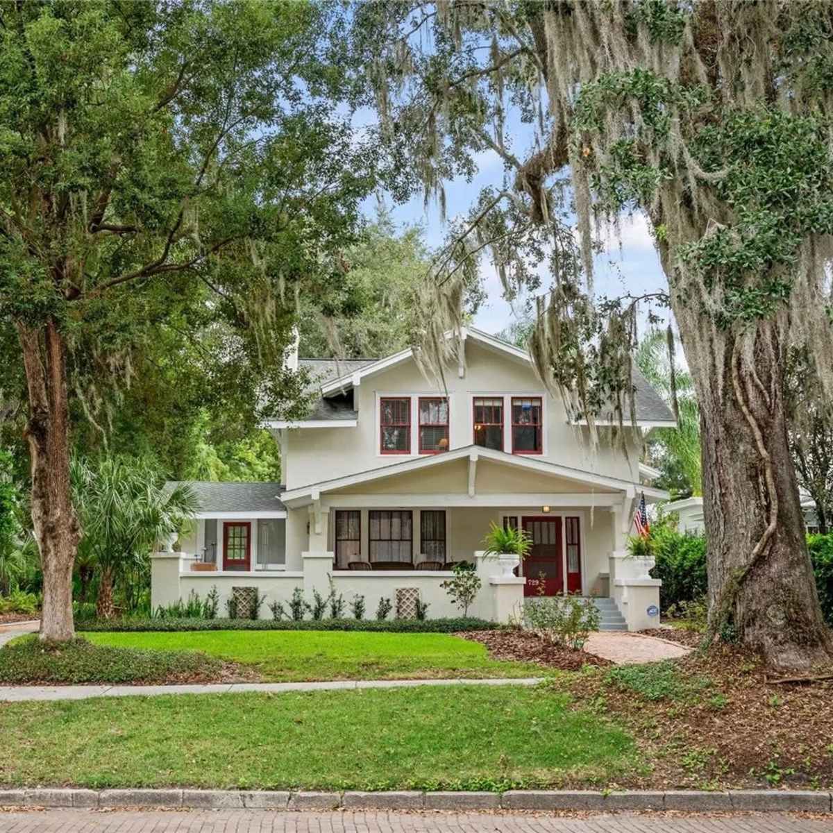 A two-story white house with red accents is surrounded by trees and landscaped greenery.