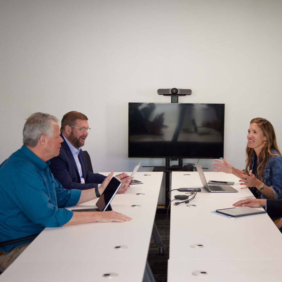 Five professionals are seated around a long white conference table engaged in a discussion, with a television monitor displaying a blurred image in the background.