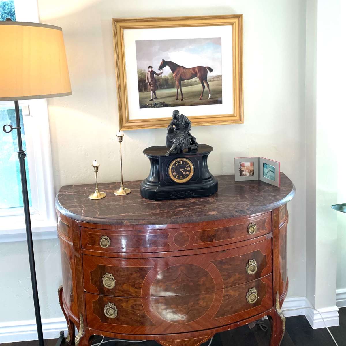 A wooden sideboard with a clock sits beneath a framed painting of a horse and rider, accompanied by a lamp and two small photo frames.