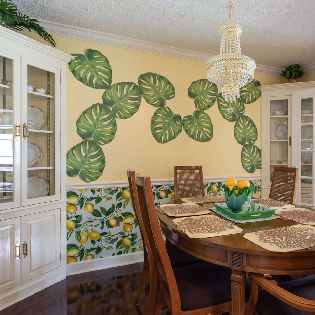 The dining room features a wooden table surrounded by chairs, with a chandelier above and a decorative mural of large leaves on the wall.