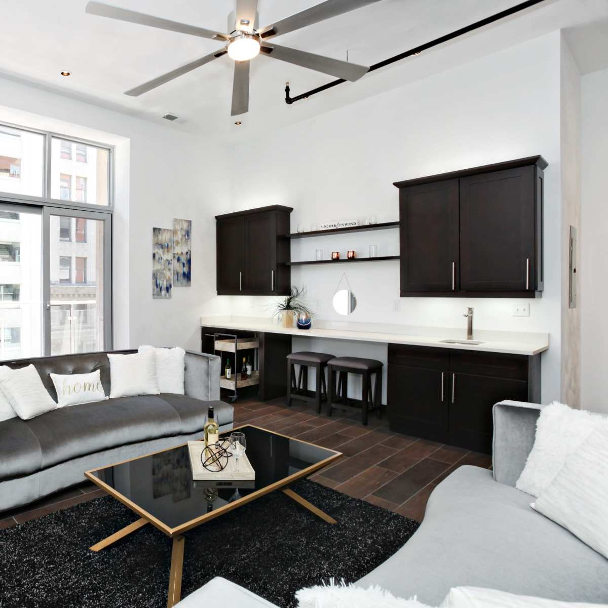 A modern living room features a gray sectional sofa, a black coffee table, and a kitchenette with dark cabinetry, illuminated by natural light from large windows.