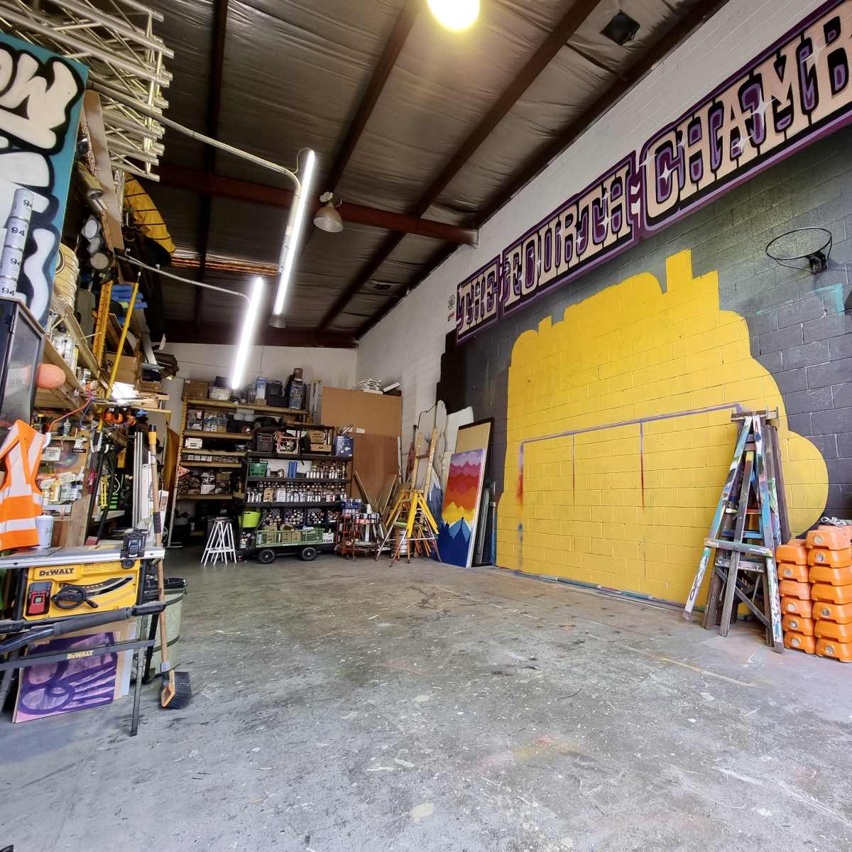 An industrial workshop space with storage racks, tools, and a partially painted wall featuring yellow and pink colors.