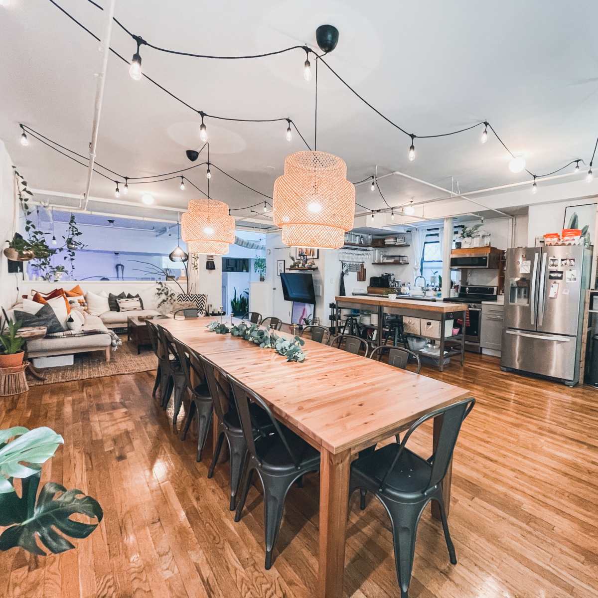A spacious dining area with a long wooden table surrounded by black chairs, pendant lights, and a cozy living space featuring a sofa and kitchen area.