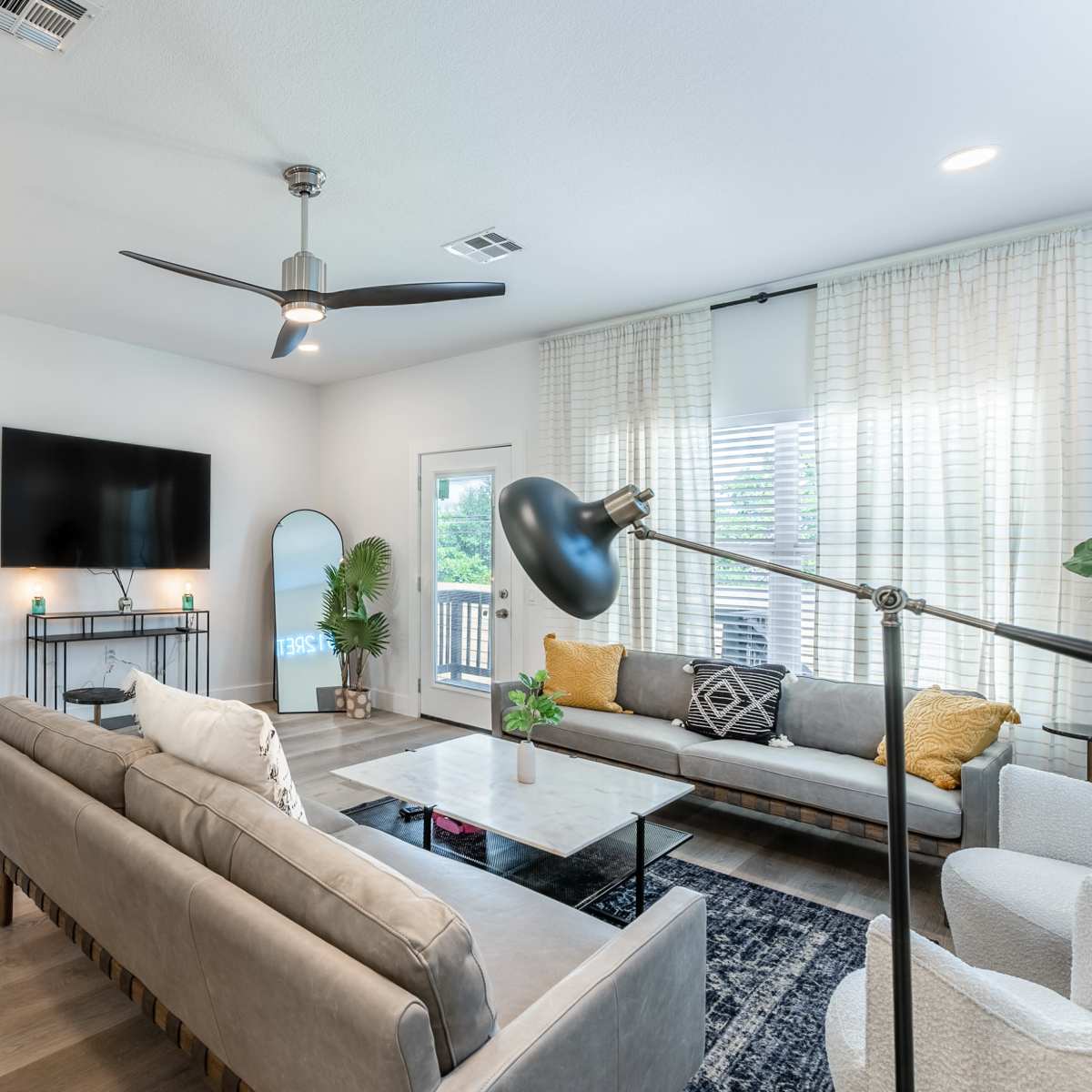 A modern living room features a gray sofa, light-colored chairs, a glass coffee table, a large wall-mounted TV, and a bar cabinet, all arranged under natural light filtering through sheer curtains.