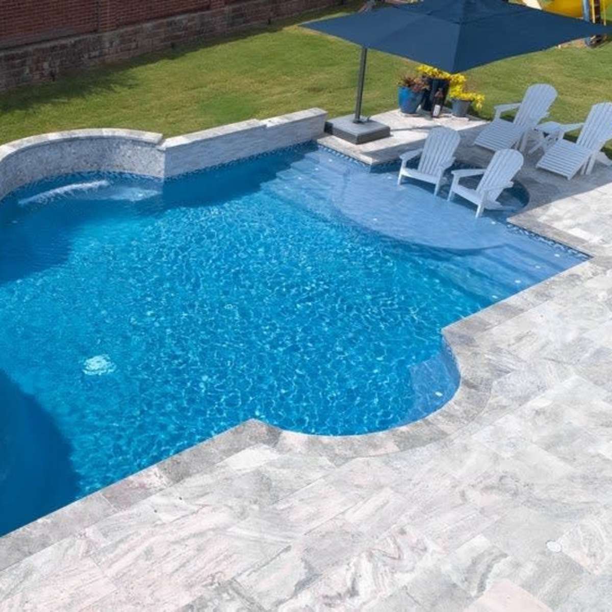 A blue swimming pool with a connected hot tub, surrounded by a patio and several white lounge chairs under an umbrella.
