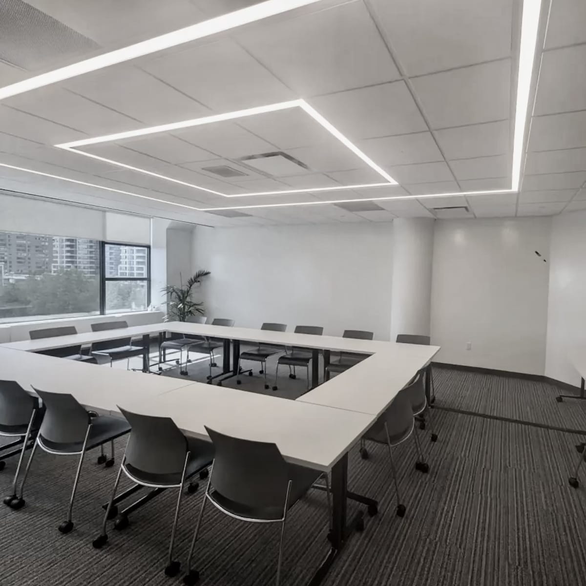A modern conference room features a long white table surrounded by grey chairs, with large windows providing natural light and a wall-mounted screen.