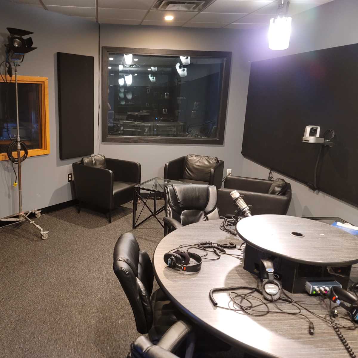 A radio studio with a round table, chairs, and sound equipment, featuring a soundproof glass window.