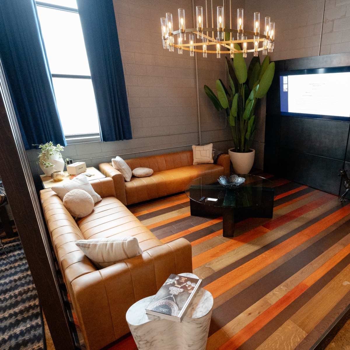 A modern lounge area featuring a brown leather sectional sofa, a black coffee table, a large potted plant, and a wall-mounted screen, all set against a backdrop of wooden striped flooring.