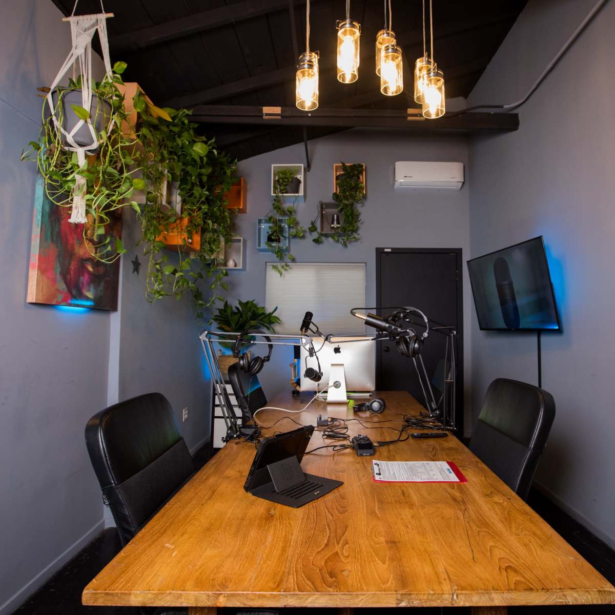 A modern office space features a wooden table with microphones and multiple monitors, surrounded by green plants and decorative wall art.
