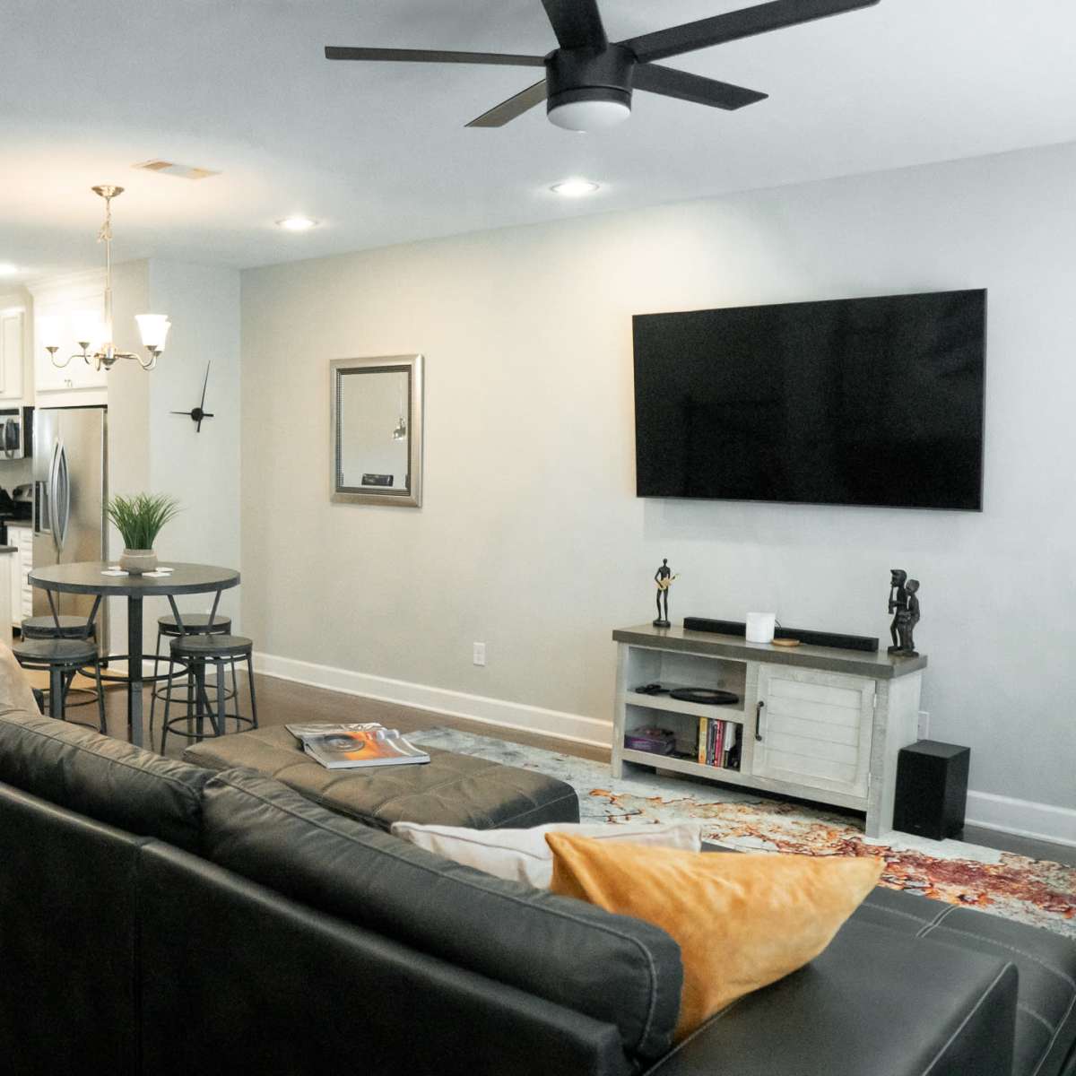 A living room with a large black sofa, a round dining table with bar stools, and an adjoining kitchen featuring white cabinets and stainless-steel appliances.