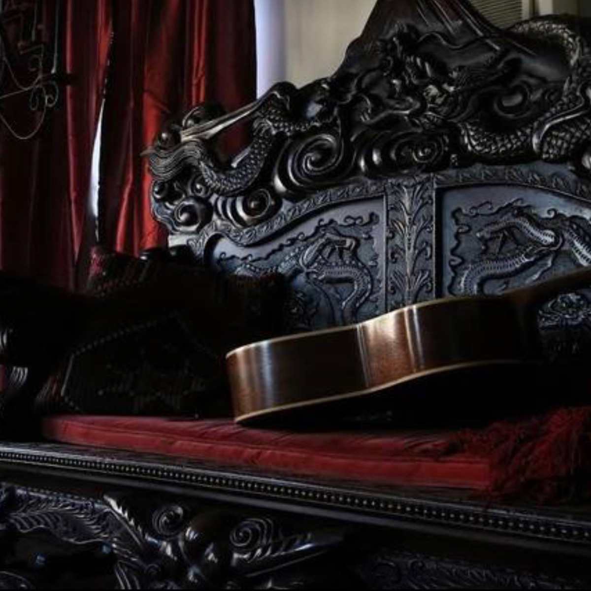A detailed, dark wooden bench with intricate carvings is adorned with a red fabric cushion and features an acoustic guitar resting against it.