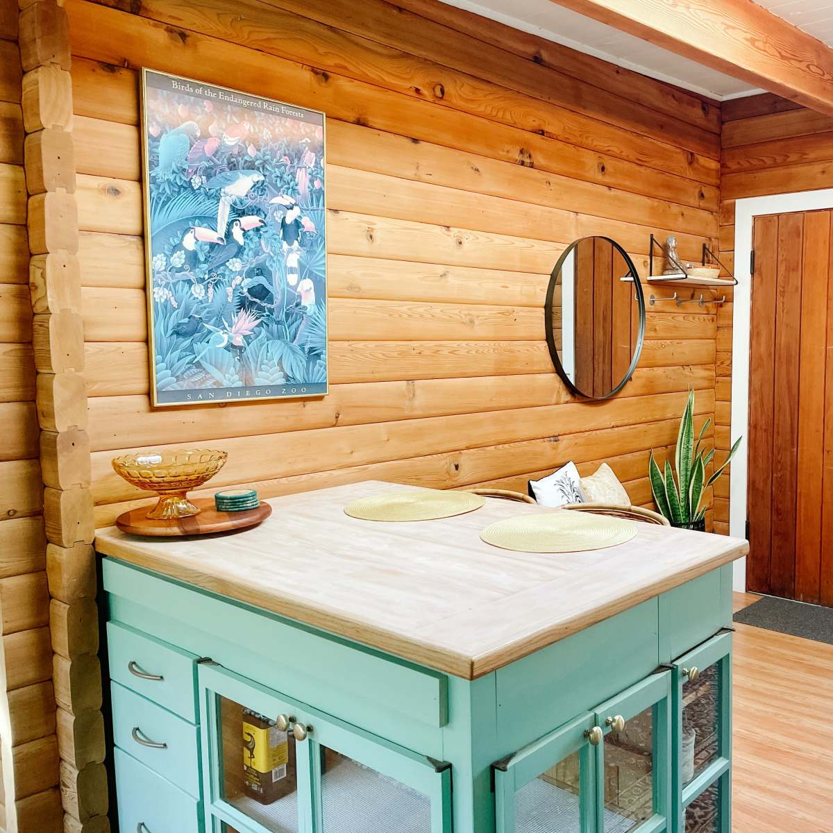 A cozy log cabin interior featuring a circular mirror, a wooden table with yellow placemats, and a doorway leading outside.