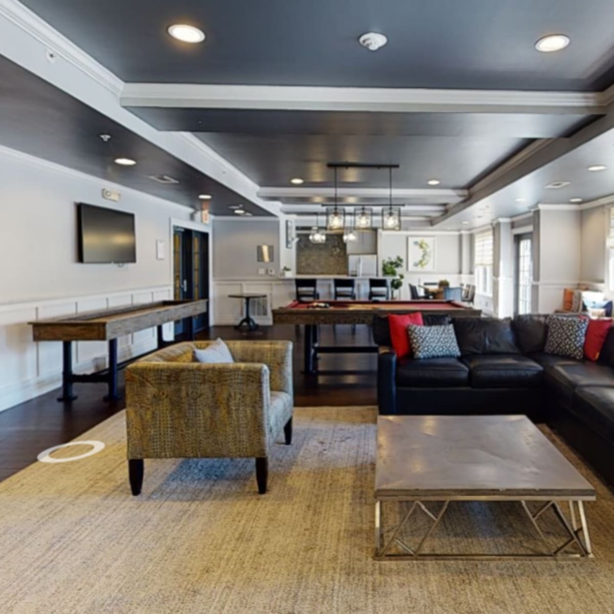 A modern lounge area featuring a dark leather sofa with red accent pillows, a coffee table, and a kitchenette in the background.