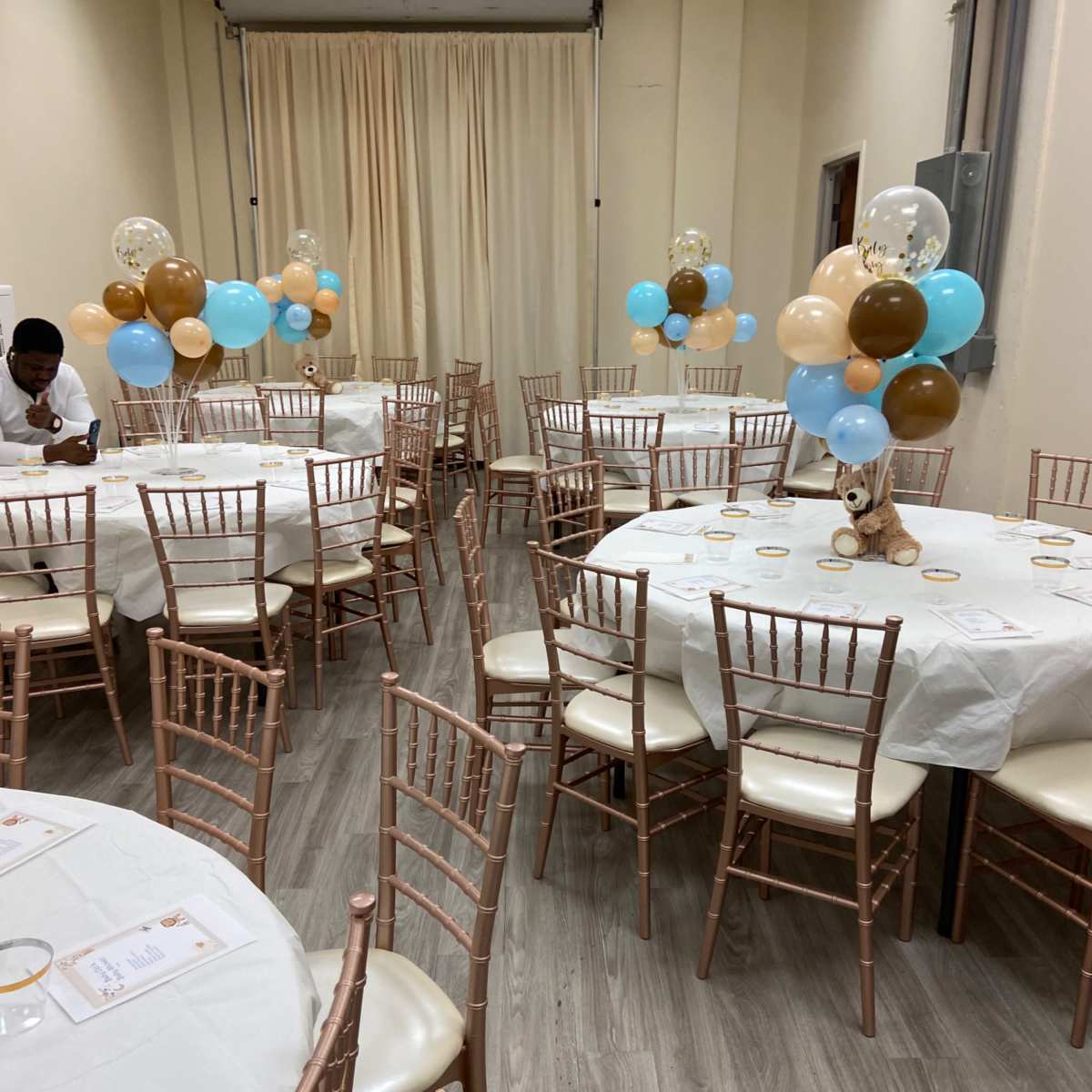 The room is set up for a celebration with white tables draped in tablecloths, surrounded by pastel balloons and rose gold chairs.