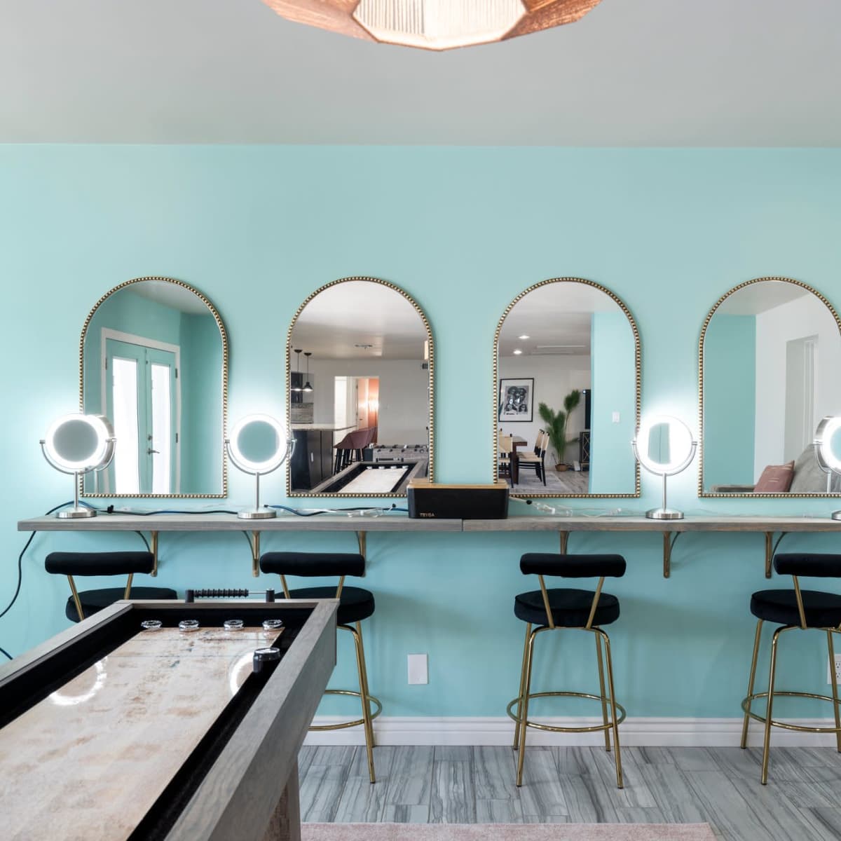 A bright room with a teal wall featuring a long table with three round mirrors and illuminated makeup stations, alongside a shuffleboard table.
