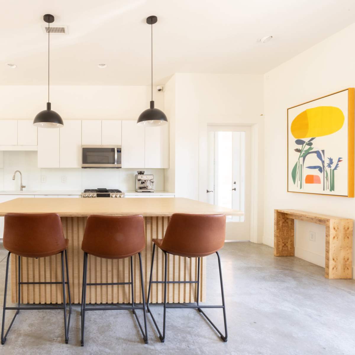 A modern kitchen features a central island with three stools, white cabinetry, stainless steel appliances, and a large abstract artwork on the wall.