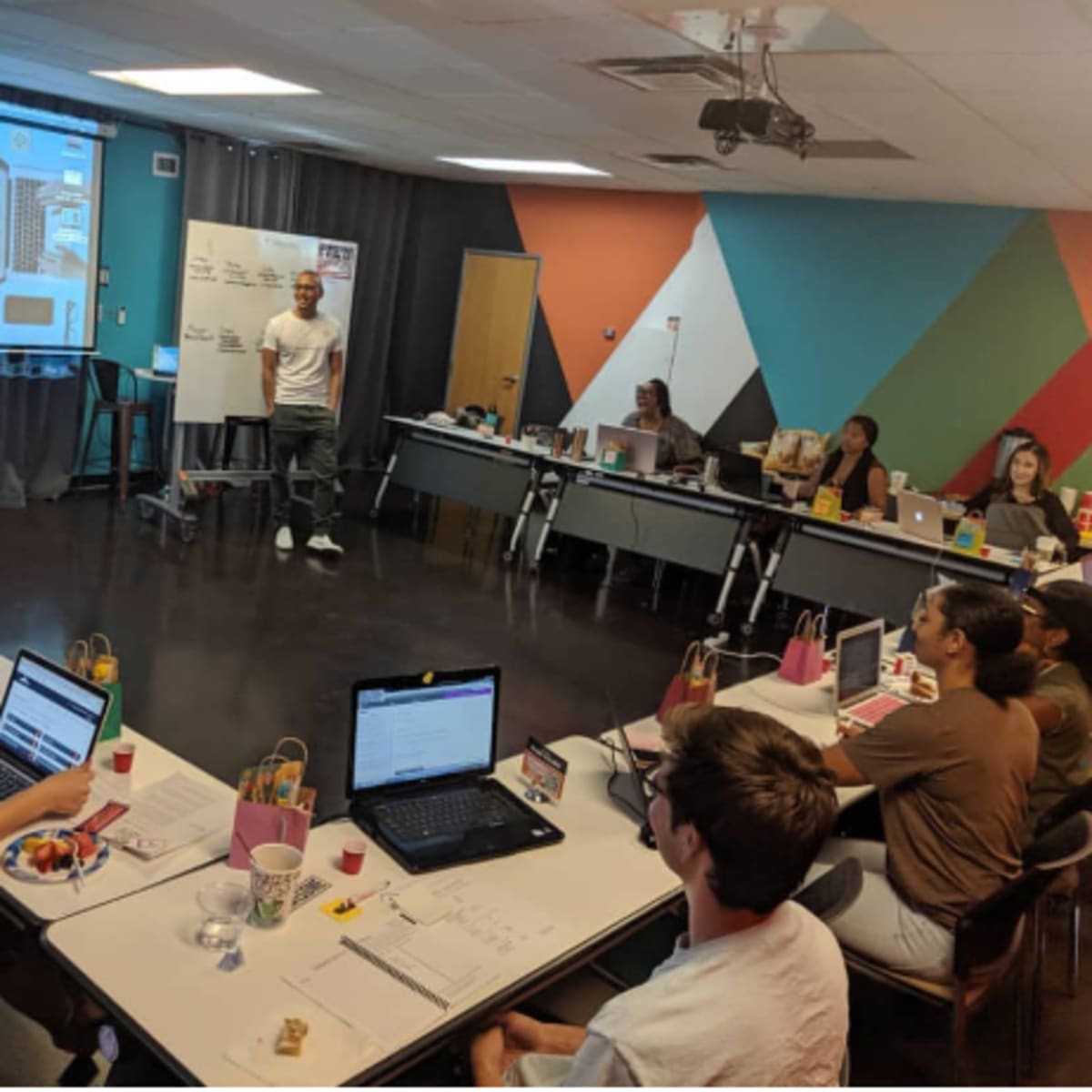 A group of people are seated at tables in a classroom setting, actively participating in a workshop titled "6-Hour Rapid Launch," with a presentation visible on a screen in the background.