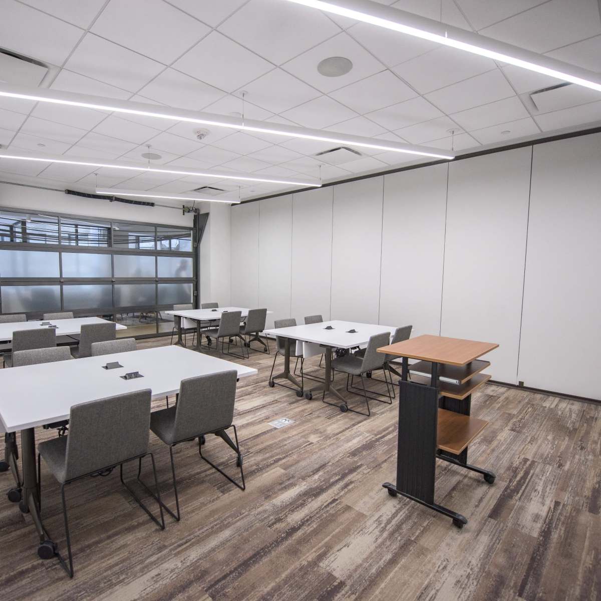 A modern classroom features multiple tables and chairs arranged for group work, with large windows and movable wall panels.
