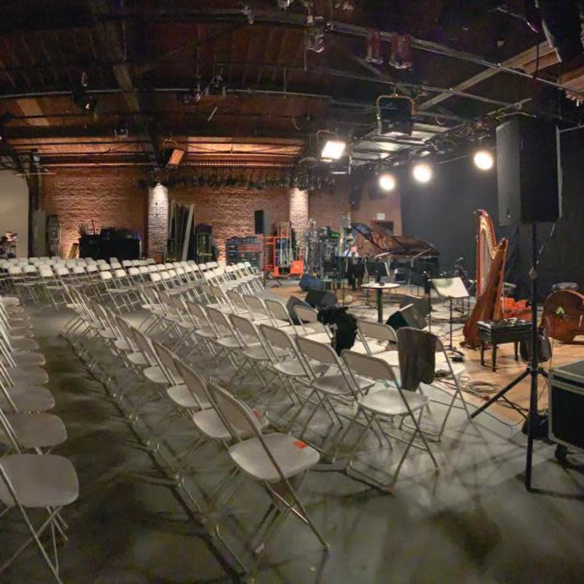 A large, empty performance space filled with rows of white folding chairs, with a stage set for a musical event in the foreground.