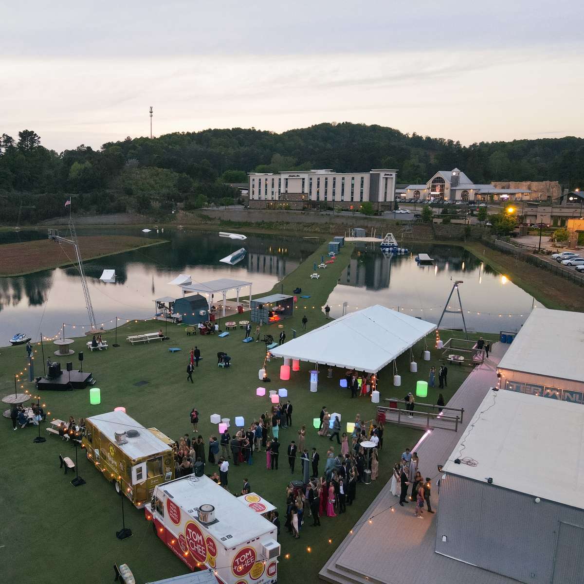 An aerial view of a lakeside event space featuring food trucks, colorful lanterns, and a gathering of people on a grassy area.