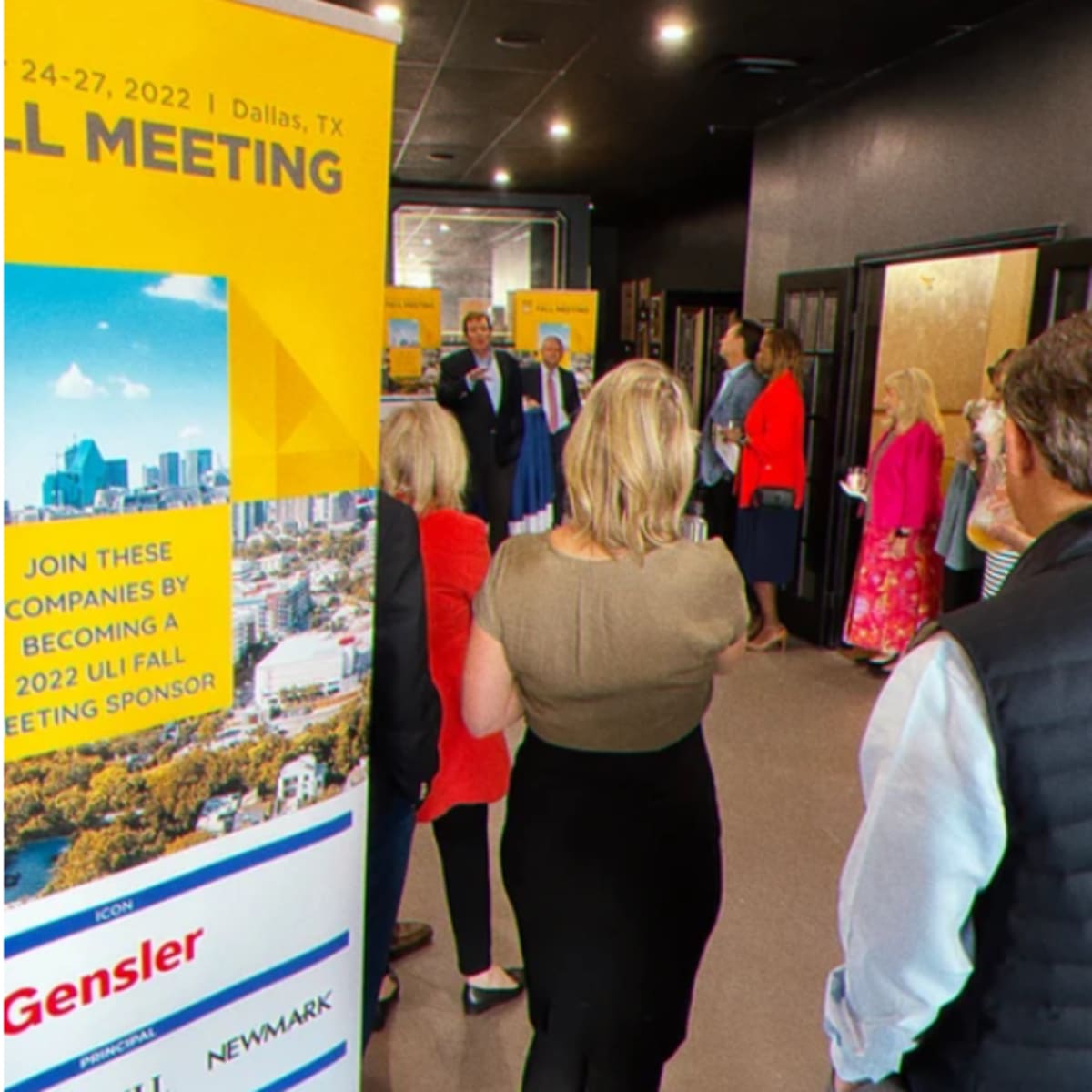 A group of people stands in a hallway attending a meeting in Dallas, Texas, with a presentation taking place in front of a large banner.