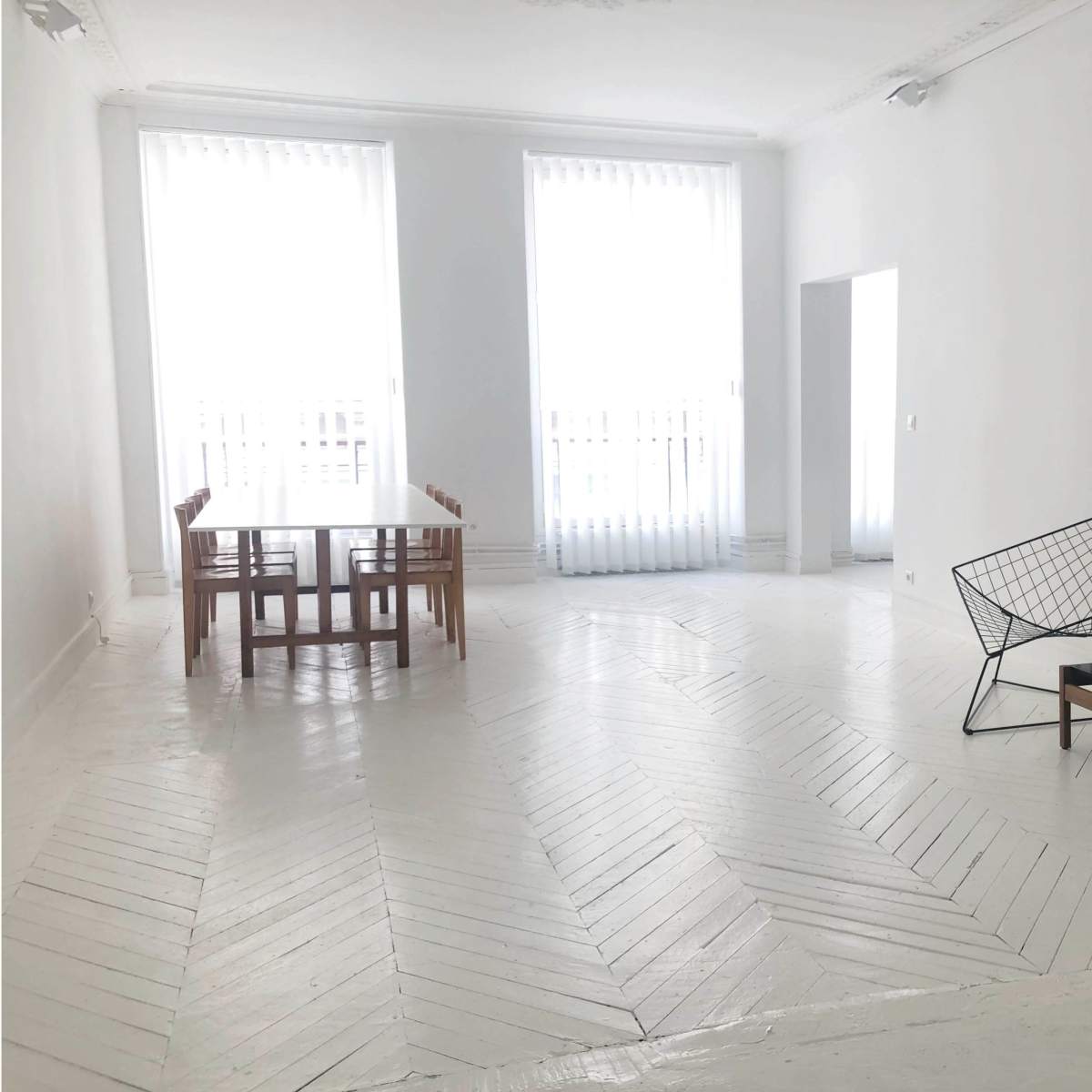 A minimalist living space features a dining table with wooden chairs, two wire-framed lounge chairs, and a lamp on a side table, all set against white walls and large windows with sheer curtains.