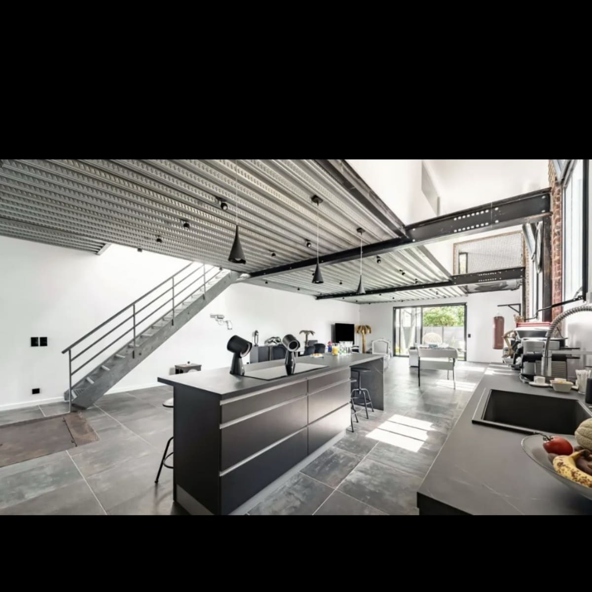 A modern, open-concept kitchen with gray cabinets and a large island, accompanied by a minimalist staircase and large windows allowing natural light to enter.