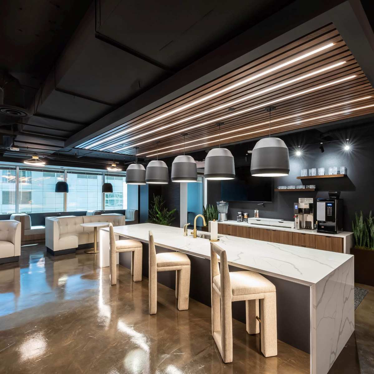 A modern office kitchen features a marble island with seating, pendant lights, and a coffee station against a backdrop of large windows.