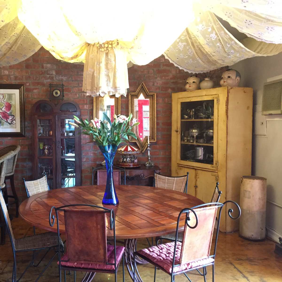 A cozy dining area with a wooden table, surrounded by various chairs, and decorated with a large draped fabric ceiling and vintage furnishings.