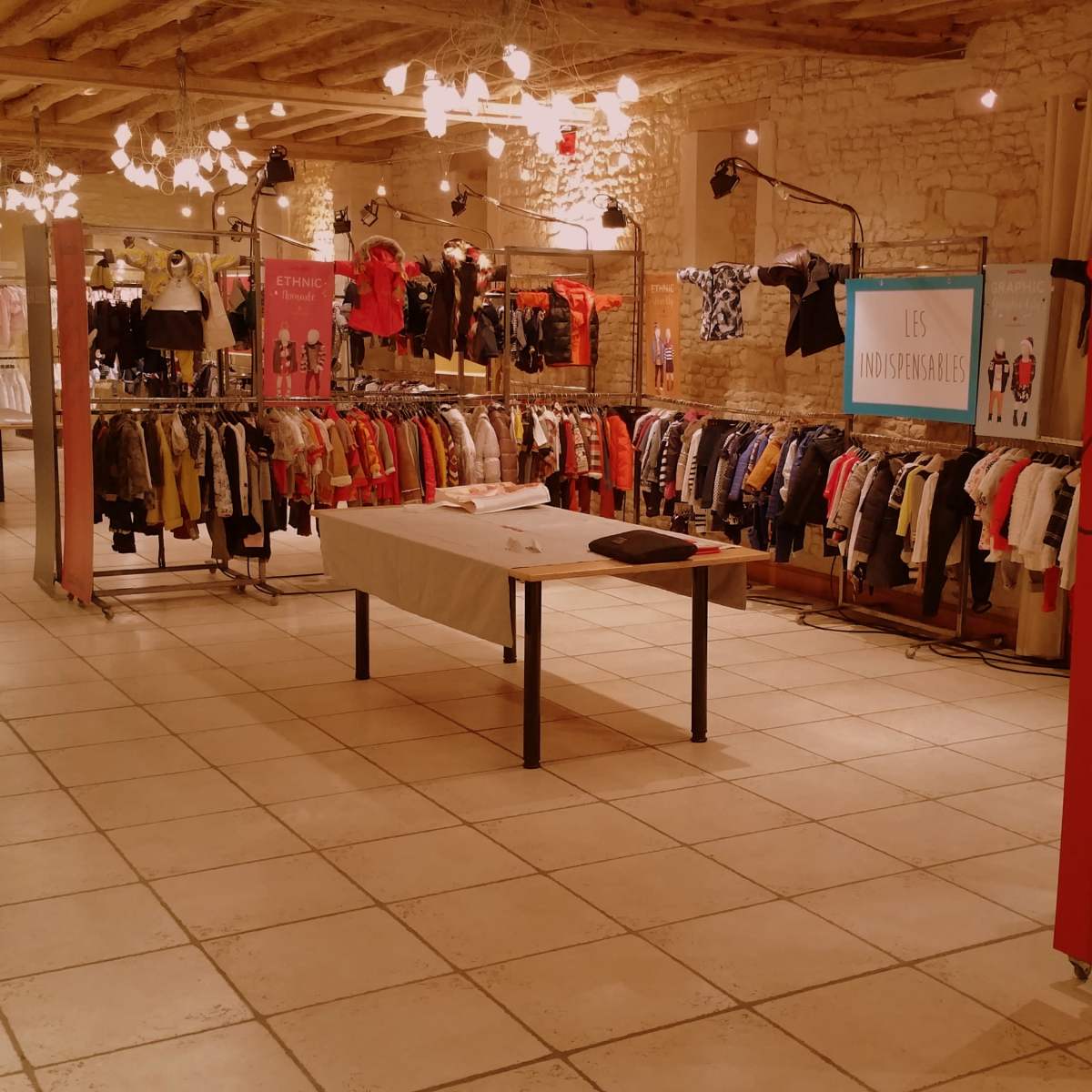 A clothing retail space featuring racks of colorful children's apparel arranged in a well-lit, spacious interior with exposed beams and decorative lighting.