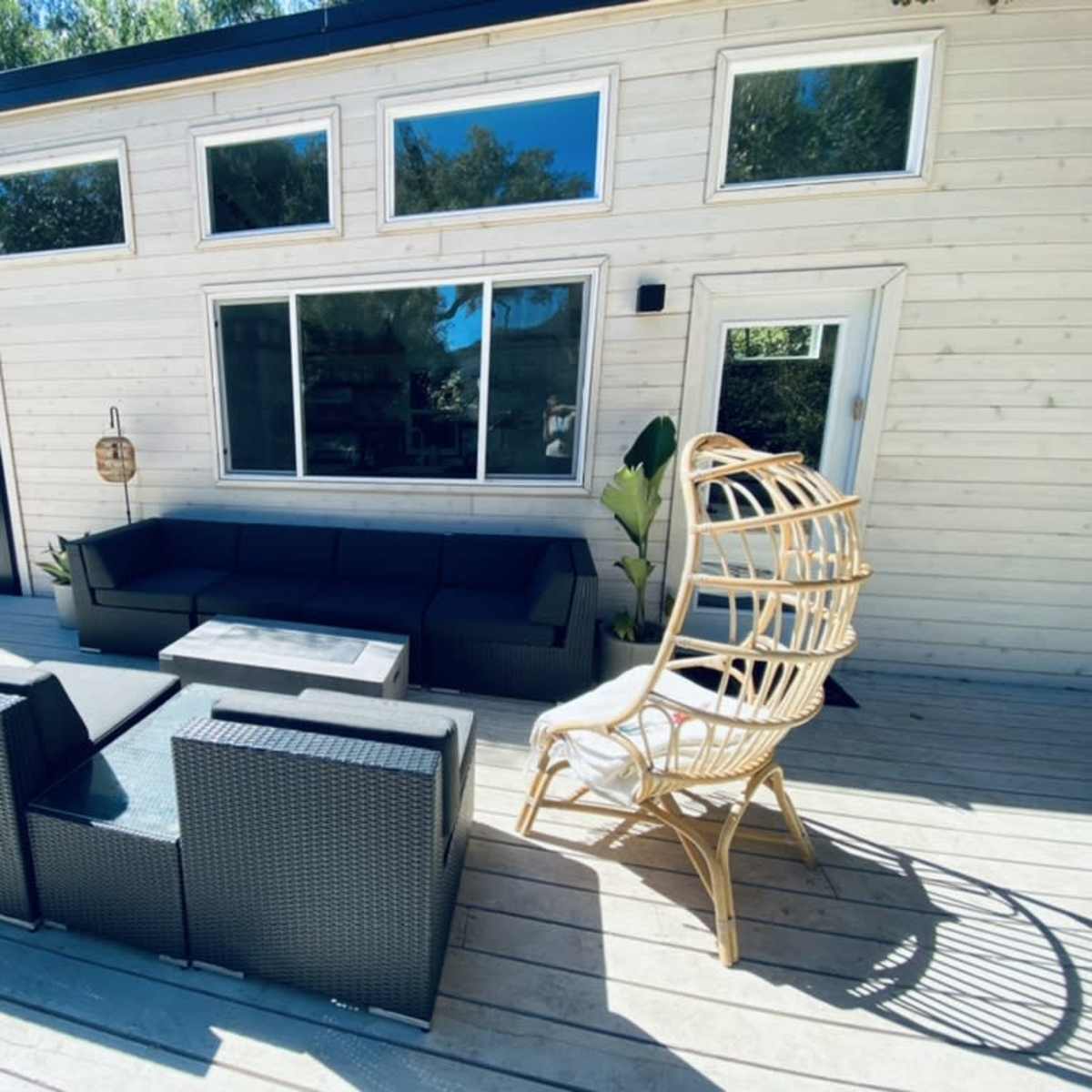 A modern wooden house with large windows, a black accent wall, a patio area with a sectional sofa, a coffee table, and a wicker chair.