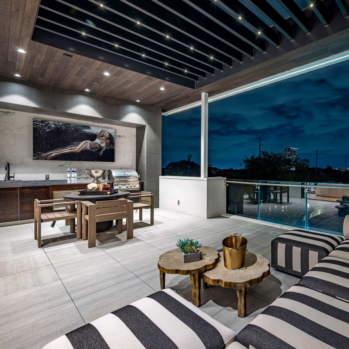 A modern outdoor patio features a striped couch, a dining area with a grill, and a city skyline visible in the background under a twilight sky.