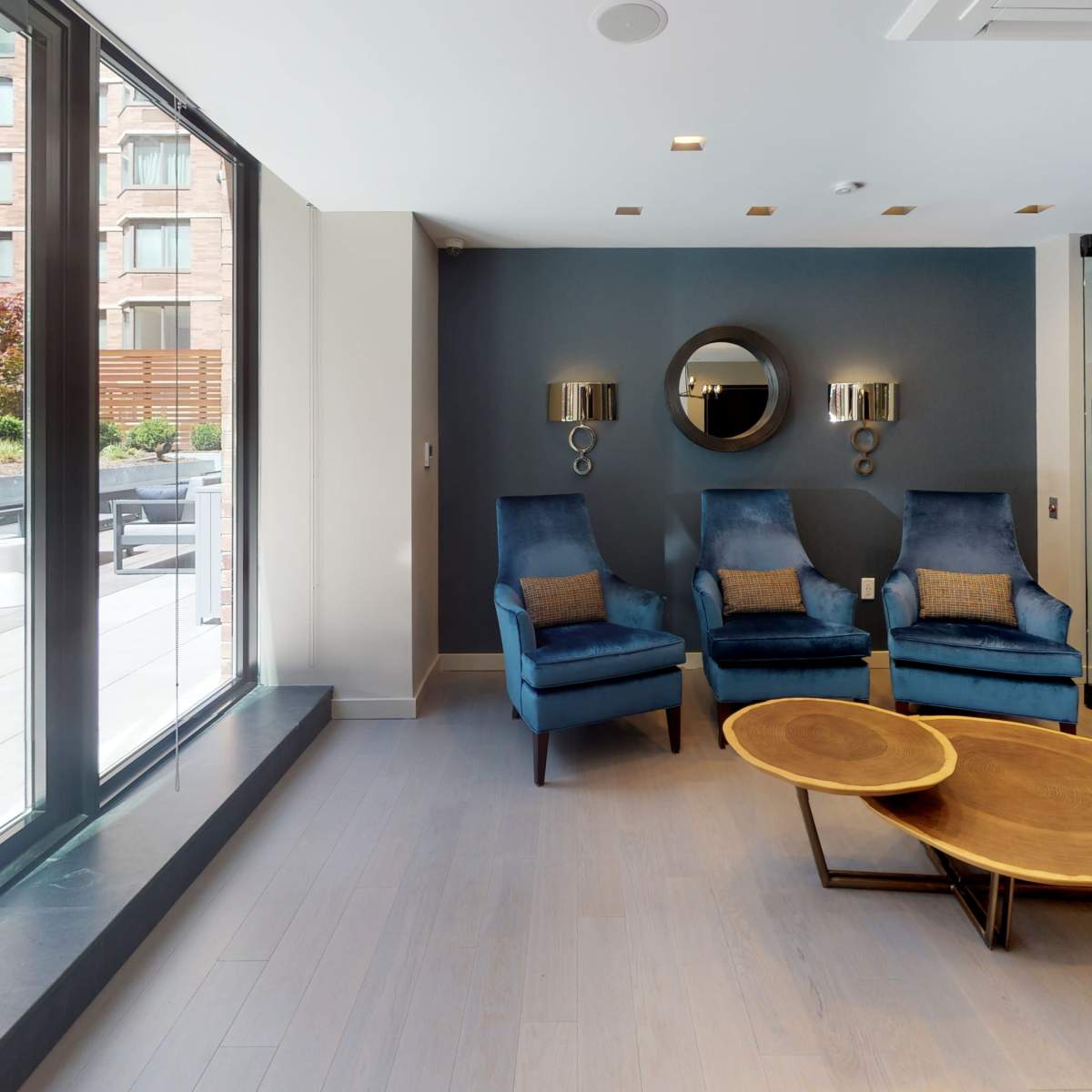 A modern lounge area featuring two blue armchairs, a round wooden coffee table, and large windows overlooking an outdoor space.
