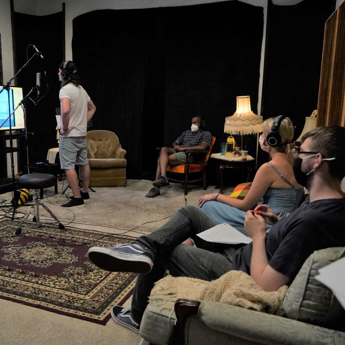 A group of four people is recording in a studio, with two seated and wearing headphones while a third stands near a microphone, watching a screen displaying animation.