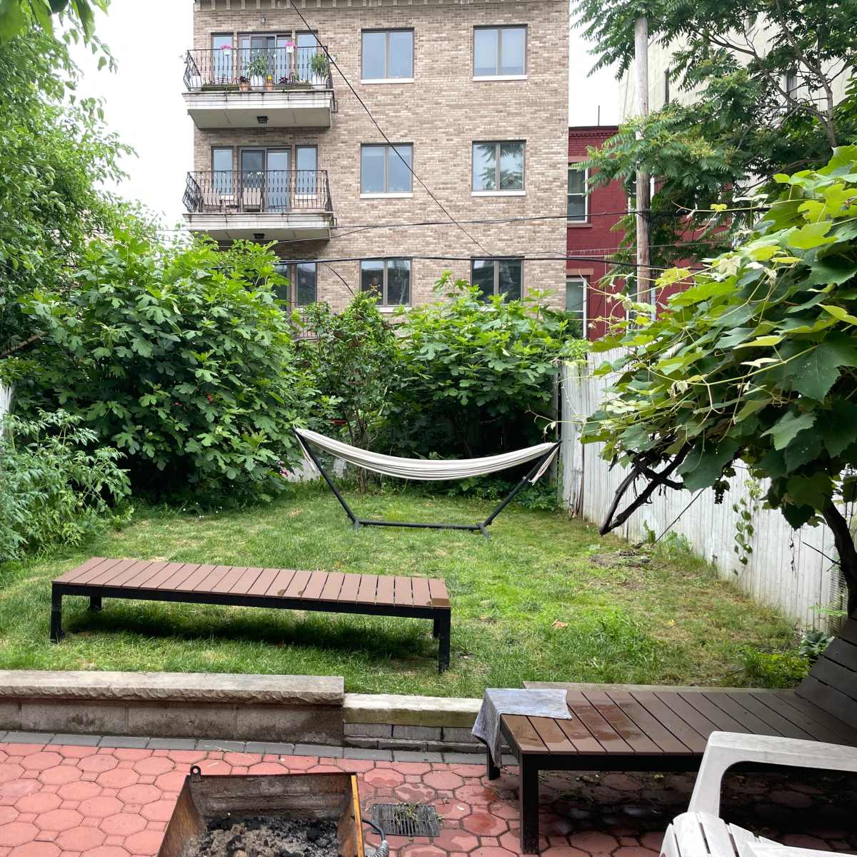 A backyard with a grassy area, a hammock between two trees, wooden lounge furniture, and a fire pit, surrounded by dense greenery and a nearby building.