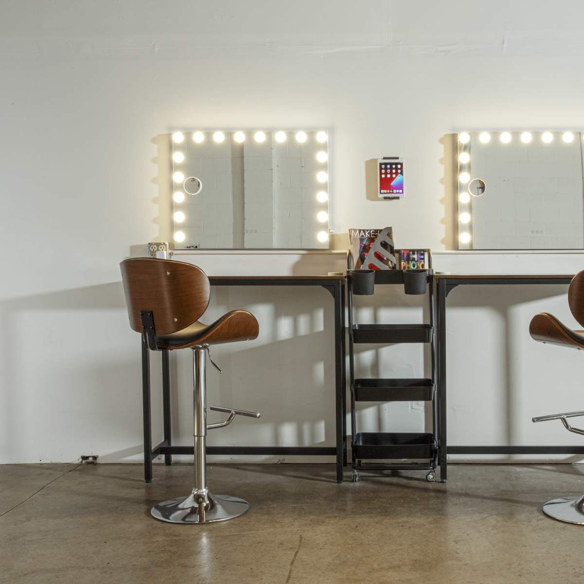 Two modern vanity tables with illuminated mirrors and high-backed stools are positioned opposite each other in a minimalist setting.