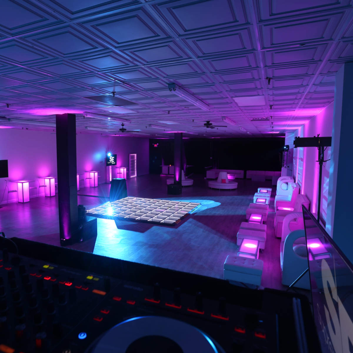 A spacious event room with a dance floor, illuminated by purple lights, featuring a DJ setup in the foreground.