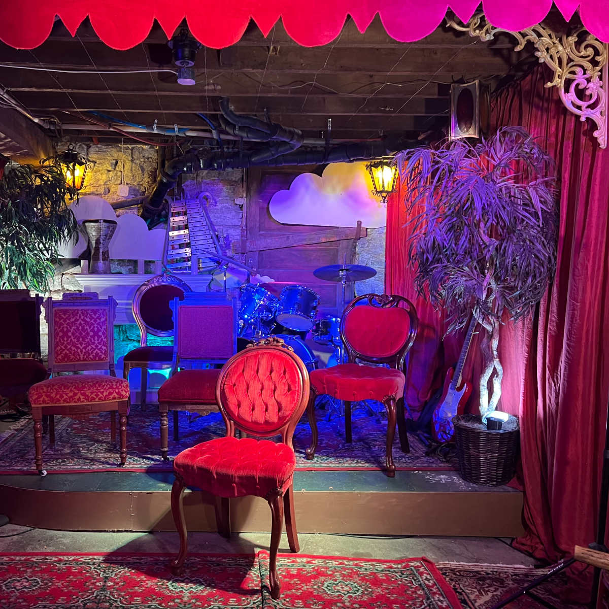The scene features a small stage adorned with various chairs, musical instruments in the background, and decorative elements such as plants and lamps.