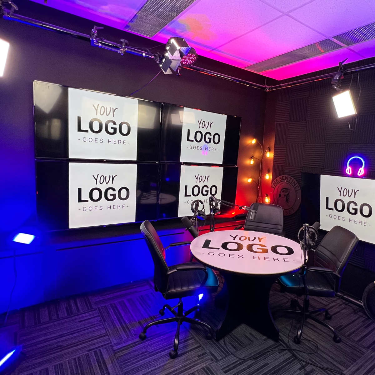 A modern studio setup for video production, featuring a round table with two chairs, multiple large screens displaying the text "YOUR LOGO GOES HERE," and various lighting fixtures.